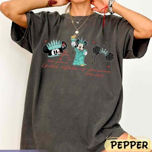 May include: Dark gray t-shirt featuring Mickey Mouse in various patriotic designs. The shirt includes Mickey Mouse wearing a Statue of Liberty crown, holding a torch, and wearing a New York hat. Text reads "250 years United States of America 1776-2026."