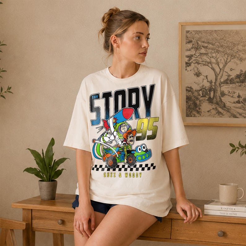 May include: Cream-colored t-shirt featuring a graphic of Buzz Lightyear and Woody in a race car, with the words "TOY STORY 95" in bold letters. The shirt has a crew neck and short sleeves. The text "BUZZ & WOODY" is printed at the bottom.