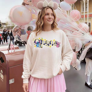 Puede incluir: Sudadera color crema con la palabra "Ohana" en un dise&ntilde;o floral, con los personajes Stitch y Angel. La persona lleva una falda plisada rosa y orejas de rat&oacute;n. Globos rosas en el fondo.