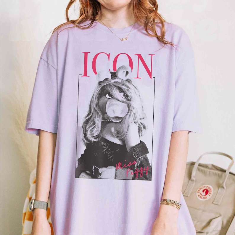 May include: A light purple t-shirt featuring a black and white image of Miss Piggy with the word "ICON" above her and "Miss Piggy" below. The shirt has short sleeves and a crew neck.