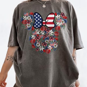 May include: Dark grey t-shirt featuring a floral design in the shape of a mouse head. The design includes a bow with the American flag and red, white, and blue flowers and flags. The shirt is a comfortable, casual style.