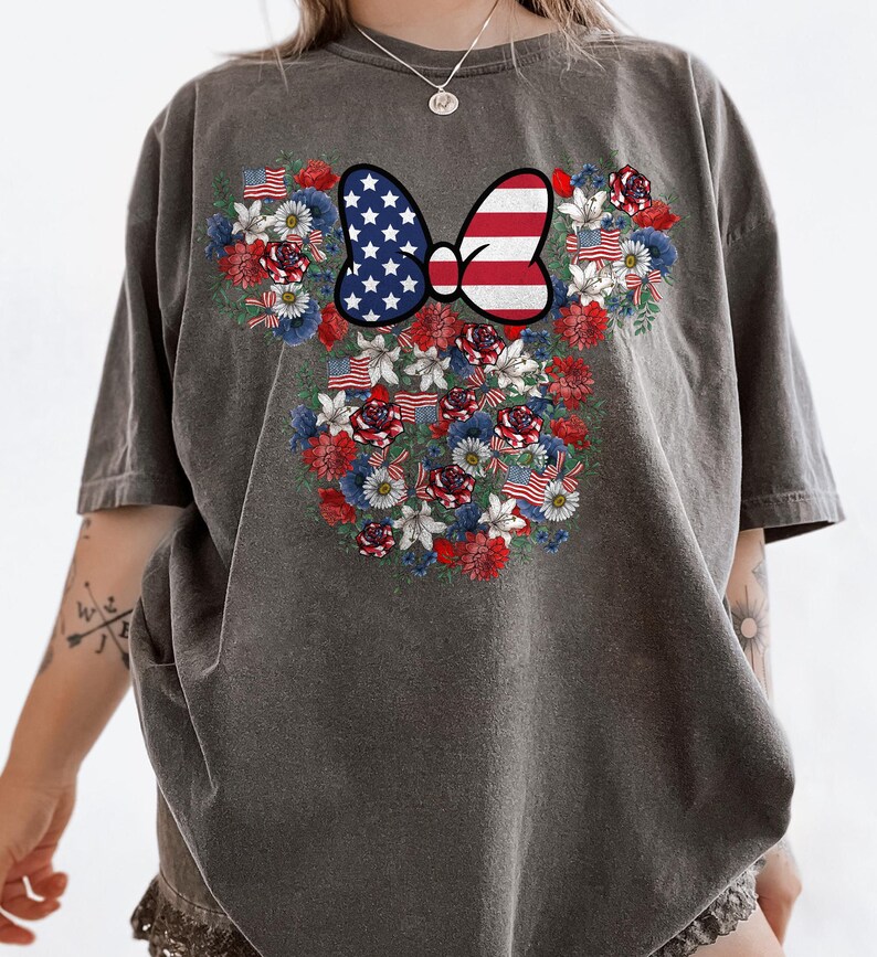 May include: Dark grey t-shirt featuring a floral design in the shape of a mouse head. The design includes a bow with the American flag and red, white, and blue flowers and flags. The shirt is a comfortable, casual style.