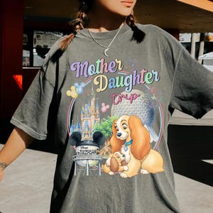 May include: Grey t-shirt featuring the text "Mother Daughter Trip" in a colourful design. The graphic includes a castle, a globe, and a cartoon dog. The shirt has short sleeves and a loose fit.