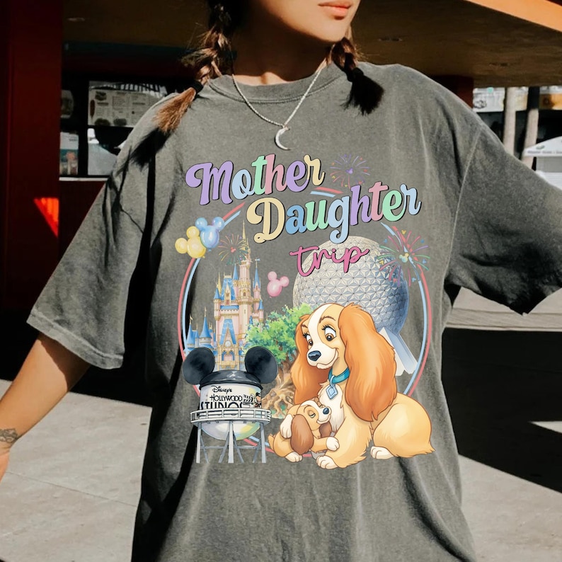 May include: Grey t-shirt featuring the text "Mother Daughter Trip" in a colourful design. The graphic includes a castle, a globe, and a cartoon dog. The shirt has short sleeves and a loose fit.