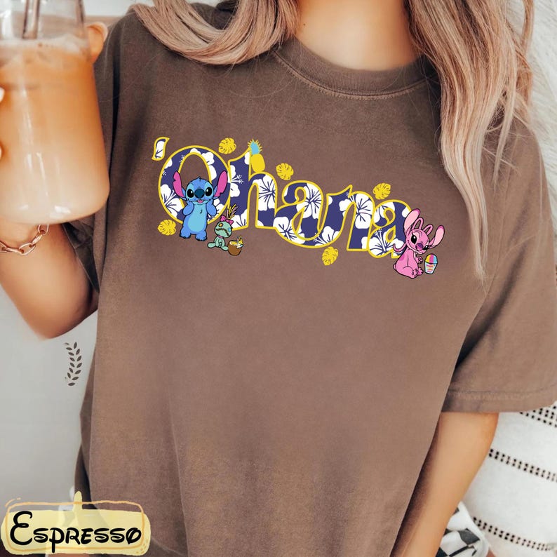 Puede incluir: Camiseta marr&oacute;n con la palabra "Ohana" en un dise&ntilde;o floral amarillo y azul. La camiseta presenta los personajes de dibujos animados Stitch y Angel, con pi&ntilde;as y flores. La camiseta es de estilo c&oacute;modo e informal.