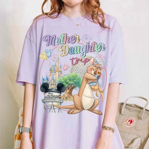 May include: A lilac t-shirt with the text "Mother Daughter Trip" in a colourful font. The design includes a cartoon kangaroo hugging a rabbit, a Disney castle, and a Hollywood Studios water tower in the background.