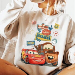 May include: Cream-coloured sweatshirt with a vibrant graphic of Lightning McQueen and Mater from the film Cars. The design includes the text "Let's Hit the Open Road" and Route 66 symbols. A playful, cartoon-style design.