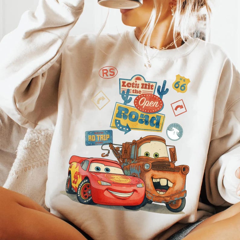 May include: Cream-coloured sweatshirt with a vibrant graphic of Lightning McQueen and Mater from the film Cars. The design includes the text "Let's Hit the Open Road" and Route 66 symbols. A playful, cartoon-style design.