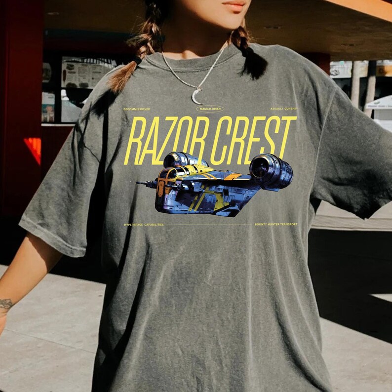 May include: Dark gray t-shirt featuring the text "RAZOR CREST" in yellow, with a detailed illustration of a spaceship. The shirt also includes text that reads "Mandalorian", "Bounty Hunter Transport", and "Apparance Capabilities".
