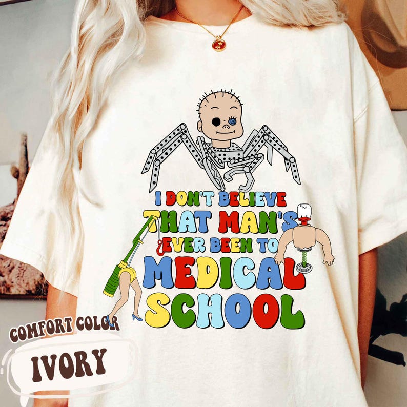 May include: Ivory-colored t-shirt with a colorful graphic. The graphic features a baby-faced spider, a dismembered torso, and a leg with a syringe. Text reads "I DON'T BELIEVE THAT MAN'S EVER BEEN TO MEDICAL SCHOOL."