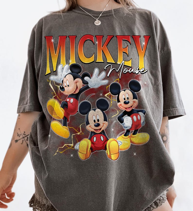 May include: Grey t-shirt featuring Mickey Mouse in three poses with yellow lightning bolts. The text "MICKEY Mouse" is printed in a gradient red and yellow font. The shirt has a vintage style.