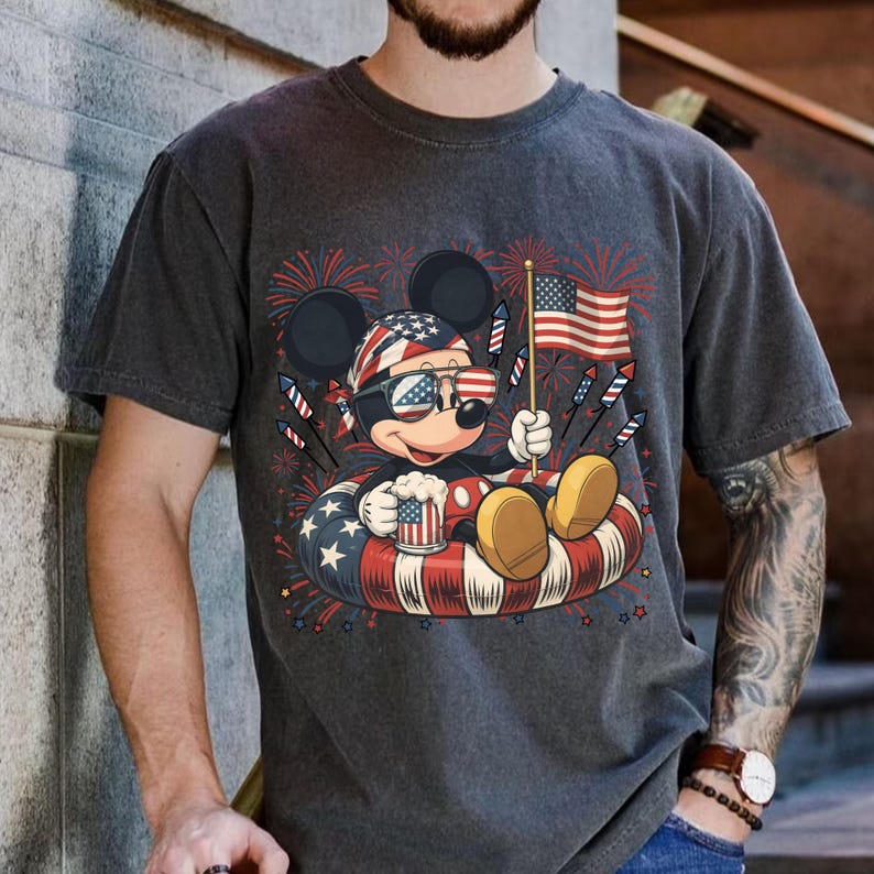 May include: Dark gray t-shirt featuring a patriotic Mickey Mouse design. Mickey is wearing sunglasses and a bandana, holding a beer mug and an American flag, surrounded by fireworks and a red, white, and blue inner tube.