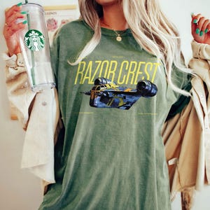 May include: Olive green t-shirt featuring the text "RAZOR CREST" in yellow, with an illustration of a spaceship. The shirt has a crew neck and short sleeves. The design is centered on the front of the shirt.
