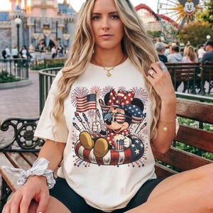 May include: Cream-colored t-shirt featuring a patriotic Minnie Mouse design. Minnie is wearing sunglasses and a bow, holding an American flag and a mug. The design includes fireworks and a red, white, and blue inner tube.