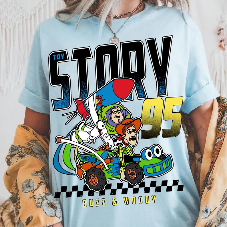 May include: Light blue t-shirt featuring a colorful graphic of Buzz Lightyear and Woody from Toy Story in a race car. The words "TOY STORY 95" are printed above the characters, with "BUZZ & WOODY" below.