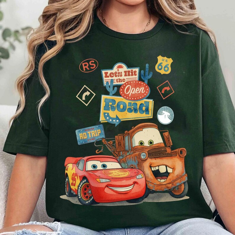 May include: Dark green t-shirt featuring Lightning McQueen and Mater from the movie Cars. The shirt has the text "Let's Hit the Open Road" with Route 66 and road trip graphics. The design is colourful and cartoon-like.
