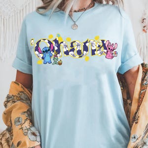 Puede incluir: Camiseta azul claro con la palabra "Ohana" en un dise&ntilde;o floral, con los personajes Stitch y Angel. La palabra "Ohana" es de color amarillo con un estampado floral azul y blanco. La camiseta tiene las mangas enrolladas.