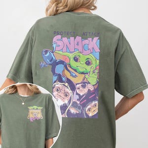 May include: Olive green t-shirt featuring a graphic of a green alien character with the text "PROTECT ATTACK SNACK". The graphic includes other characters in a purple and blue design. A smaller version of the graphic is on the front.