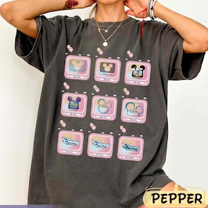 Puede incluir: Camiseta gris oscuro con una cuadrícula de televisores rosas que muestran logotipos de Disney Channel e imágenes de Mickey Mouse. La camiseta tiene un corte holgado y un aspecto vintage, con la palabra "PEPPER" en un rectángulo amarillo.