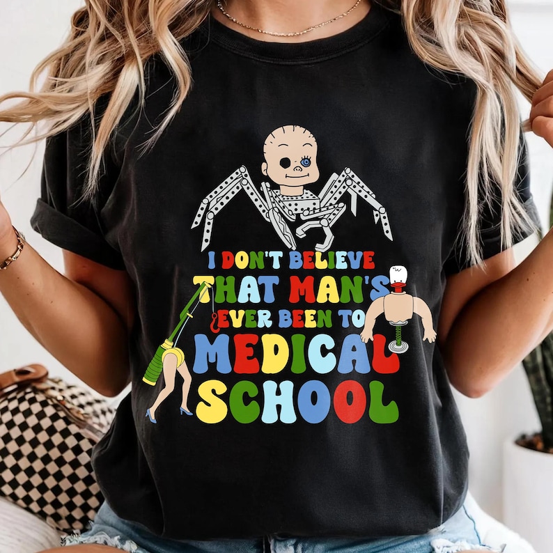 May include: Black t-shirt with a humorous graphic. The design features a baby-faced spider, a figure with a medical instrument, and the text "I DON'T BELIEVE THAT MAN'S EVER BEEN TO MEDICAL SCHOOL" in colorful letters.