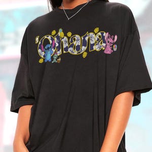 Puede incluir: Camiseta negra con la palabra "Ohana" en un dise&ntilde;o tropical colorido. La palabra est&aacute; delineada en amarillo y rellena con un estampado floral. Tambi&eacute;n aparecen los personajes de dibujos animados Stitch y Angel.