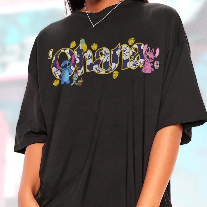 Puede incluir: Camiseta negra con la palabra "Ohana" en un dise&ntilde;o tropical colorido. La palabra est&aacute; delineada en amarillo y rellena con un estampado floral. Tambi&eacute;n aparecen los personajes de dibujos animados Stitch y Angel.