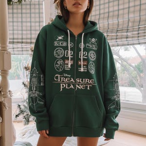 May include: A dark green zip-up hoodie featuring the text "Disney Treasure Planet" in white. The hoodie has various white graphic patches and a ship blueprint design on the sleeves. It has a drawstring hood and a front pocket.
