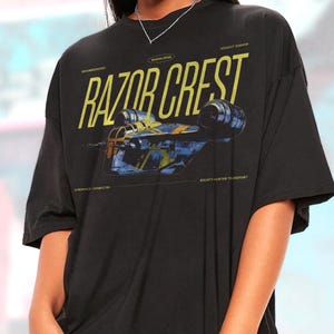 May include: Black t-shirt featuring a yellow and blue graphic of the Razor Crest spaceship. The text "RAZOR CREST" is prominently displayed above the image, with additional text indicating "Mandalorian", "Assault Gunship", "Hyperspace Capabilities", and "Bounty Hunter Transport".