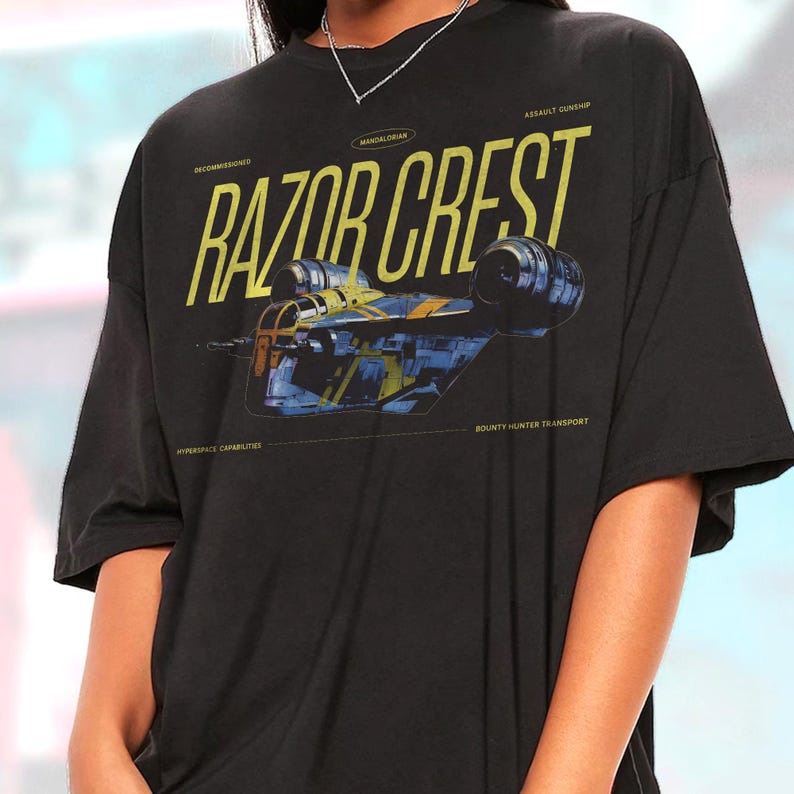 May include: Black t-shirt featuring a yellow and blue graphic of the Razor Crest spaceship. The text "RAZOR CREST" is prominently displayed above the image, with additional text indicating "Mandalorian", "Assault Gunship", "Hyperspace Capabilities", and "Bounty Hunter Transport".
