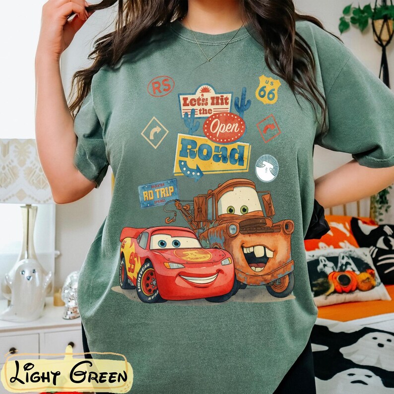 May include: Light green t-shirt featuring Lightning McQueen and Mater from the movie Cars, with the text "Let's Hit the Open Road." The design includes road trip-themed graphics and the text "Light Green" at the bottom.