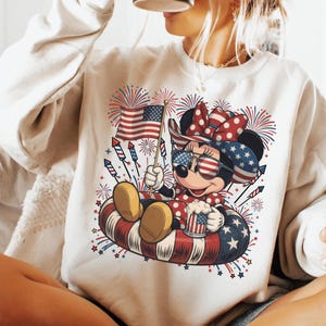 May include: Cream-colored sweatshirt featuring a patriotic Minnie Mouse design. Minnie is wearing sunglasses, a bow, and a polka-dot dress, holding an American flag and a mug. Fireworks and stars surround her, with a red, white, and blue color scheme.