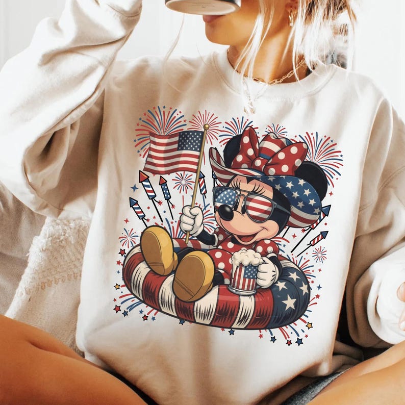 May include: Cream-colored sweatshirt featuring a patriotic Minnie Mouse design. Minnie is wearing sunglasses, a bow, and a polka-dot dress, holding an American flag and a mug. Fireworks and stars surround her, with a red, white, and blue color scheme.