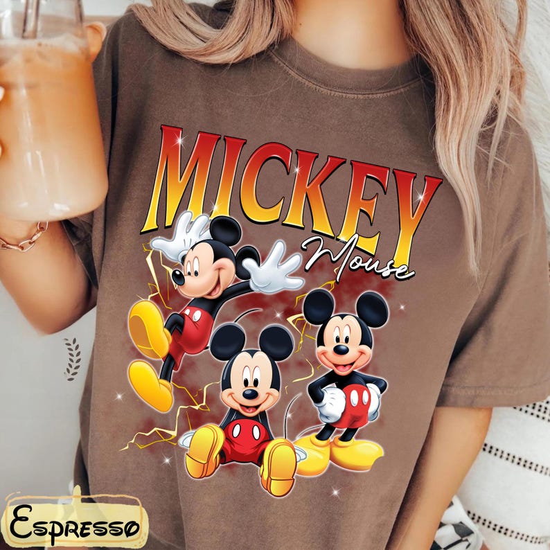 May include: Brown t-shirt with a graphic of Mickey Mouse in three poses. The text "Mickey Mouse" is in yellow and red. Mickey is wearing red shorts, yellow shoes, and white gloves. A fun, cartoon-themed top.