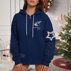 May include: A navy blue zip-up hoodie featuring a Mickey Mouse Fantasia design on the chest and star graphics on the sleeve. The hoodie has a drawstring hood, front pockets, and the text "85th" on the bottom.