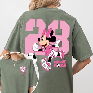 May include: An olive green t-shirt with a pink graphic of Minnie Mouse playing football, with the number 23 in pink. The words "MINNIE MOUSE" are printed below the character. A smaller image of the design is on the front.