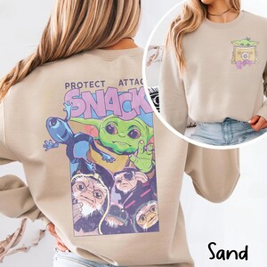 May include: Sand-colored sweatshirt featuring a colorful graphic of a green alien character with the text "PROTECT SNACK ATTAC". The design includes other alien characters. A smaller graphic of the alien character is on the front.