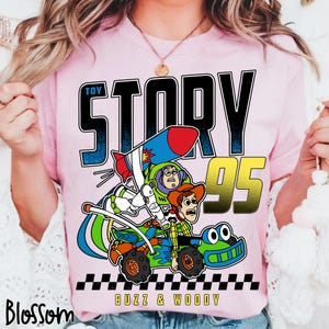May include: Pink t-shirt featuring a graphic of Buzz Lightyear and Woody from Toy Story in a race car. The text "TOY STORY" is above the image, with "95" in gold and "BUZZ & WOODY" below. A black and white checkered pattern is at the bottom.