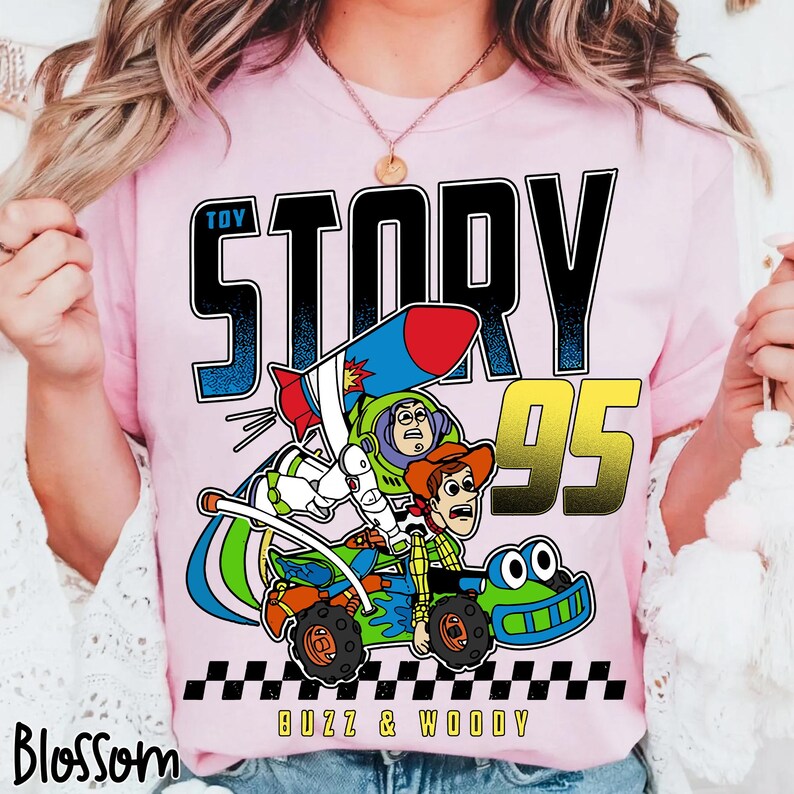 May include: Pink t-shirt featuring a graphic of Buzz Lightyear and Woody from Toy Story in a race car. The text "TOY STORY" is above the image, with "95" in gold and "BUZZ & WOODY" below. A black and white checkered pattern is at the bottom.
