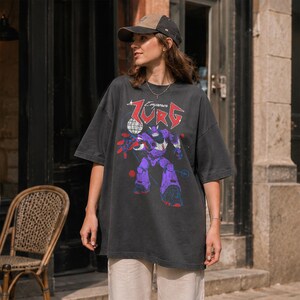 Puede incluir: Camiseta gris oscuro de gran tama&ntilde;o con un gr&aacute;fico de robot p&uacute;rpura y el texto "Zurg Emperor" en rojo y blanco. La camiseta se usa con una gorra de b&eacute;isbol marr&oacute;n y beige.