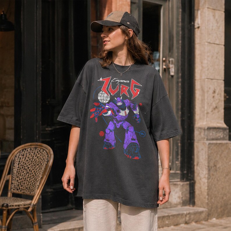Puede incluir: Camiseta gris oscuro de gran tama&ntilde;o con un gr&aacute;fico de robot p&uacute;rpura y el texto "Zurg Emperor" en rojo y blanco. La camiseta se usa con una gorra de b&eacute;isbol marr&oacute;n y beige.