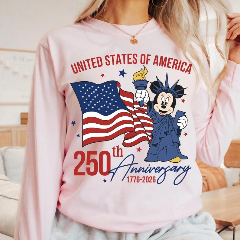 May include: A light pink long-sleeved shirt featuring a graphic celebrating the 250th anniversary of the United States of America. The design includes the American flag, a cartoon character dressed as the Statue of Liberty, and the text "United States of America" and "250th Anniversary 1776-2026".