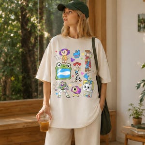 May include: Cream-colored oversized t-shirt featuring colorful cartoon character graphics. The design includes images of a frog, a robot, a cowboy, and other characters. The shirt is a casual, comfortable style.