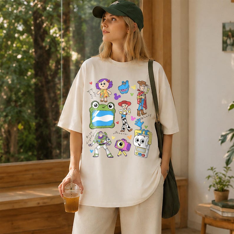 May include: Cream-colored oversized t-shirt featuring colorful cartoon character graphics. The design includes images of a frog, a robot, a cowboy, and other characters. The shirt is a casual, comfortable style.