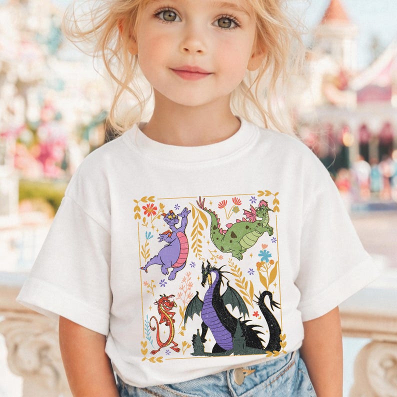 May include: White t-shirt featuring a colorful graphic of cartoon dragons. The design includes a purple dragon, a green dragon, a black dragon, and a red dragon, surrounded by flowers and leaves. The shirt is worn by a young child.