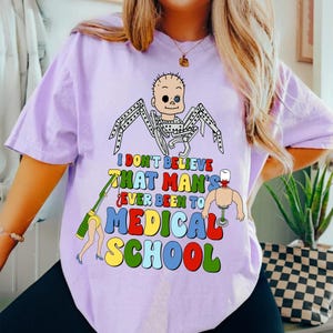 May include: A light purple t-shirt with a colorful graphic that reads "I don't believe that man's ever been to medical school." The graphic features a cartoon baby with spider legs and other medical-themed illustrations.