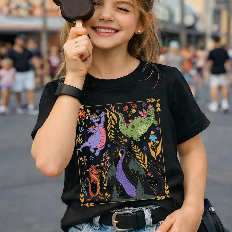 May include: Black t-shirt featuring a colorful graphic of cartoon dragons and floral elements within a gold border. The design includes a purple, green, red, and black dragon. The shirt is worn by a person holding a chocolate ice cream bar.
