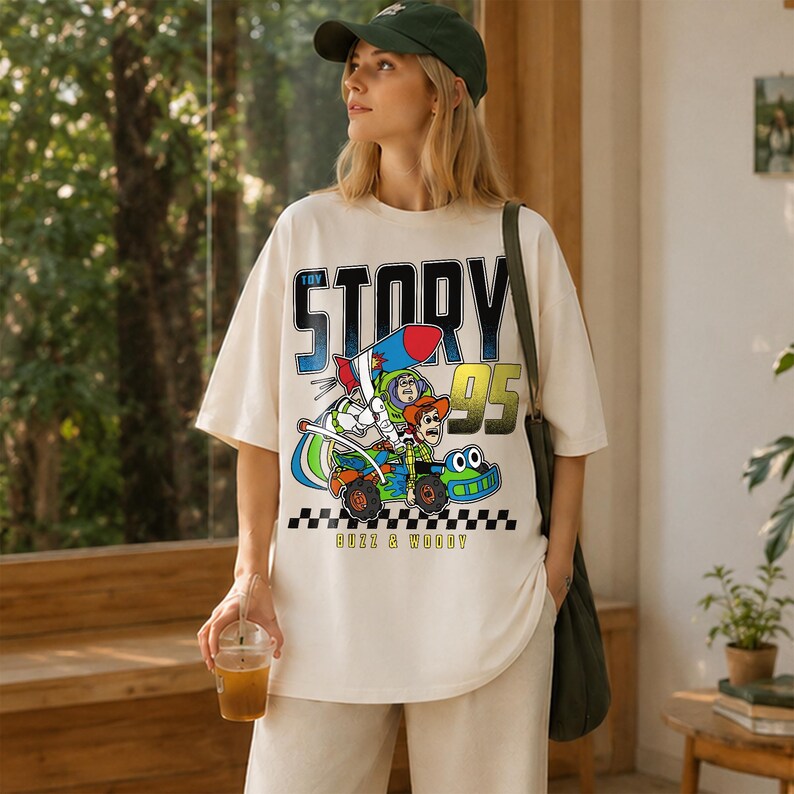 May include: Cream-colored t-shirt featuring a colorful graphic of Buzz Lightyear and Woody from Toy Story in a race car. The shirt has the words "Toy Story 95" printed on it, with "Buzz & Woody" below the car. The shirt is a relaxed fit.