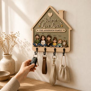 May include: A house-shaped wooden wall key holder with a sage green background, featuring the text Where Love Lives and THE ANDERSON FAMILY. It includes five small figurines of people and a cat, with four black metal hooks holding various keychains.