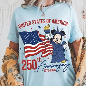 May include: Light blue t-shirt with the text "UNITED STATES OF AMERICA" in red. The shirt features a waving American flag and a cartoon character dressed as the Statue of Liberty. The shirt also has the text "250th Anniversary 1776-2026".