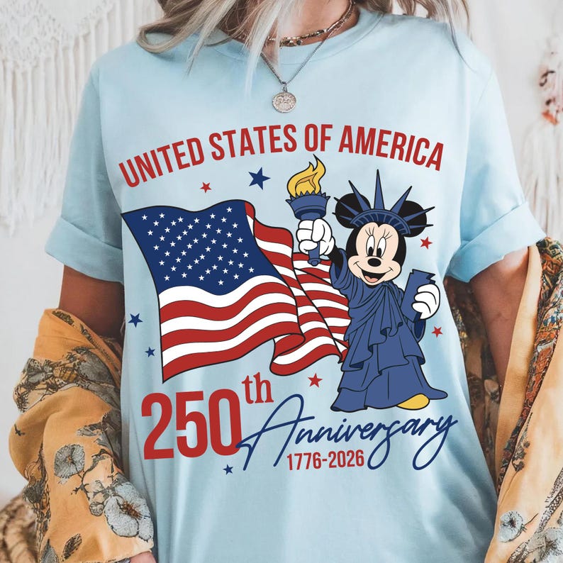 May include: Light blue t-shirt with the text "UNITED STATES OF AMERICA" in red. The shirt features a waving American flag and a cartoon character dressed as the Statue of Liberty. The shirt also has the text "250th Anniversary 1776-2026".